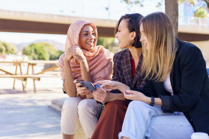Three happy women sitting on outdoor park bench watching smartphone