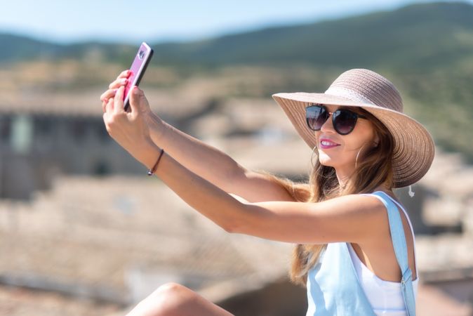 Positive woman in hat taking selfie on smartphone while standing on street