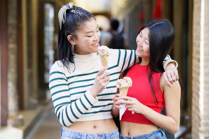 Cheerful Asian female standing with hand on shoulder of friend while eating delicious ice cream in street