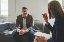 A white man in his 40s, dressed in a jacket and shirt, sits on his therapist's couch with interlocked finger and downcast eyes