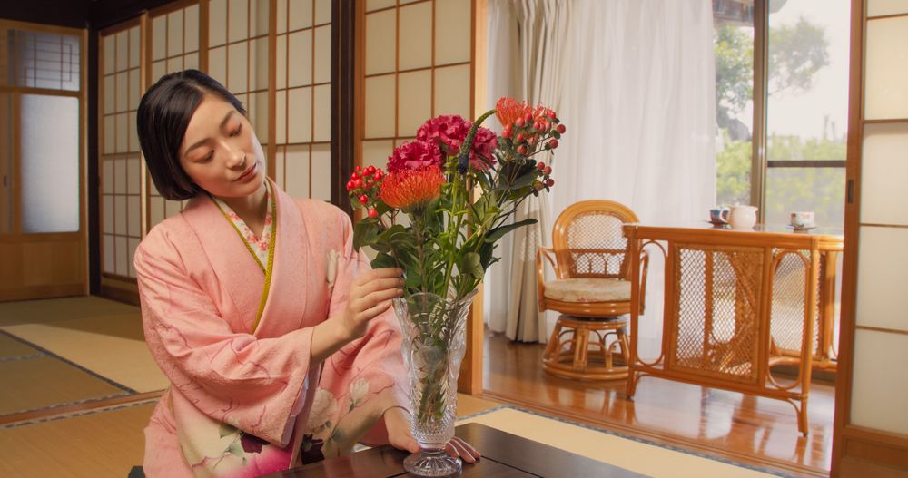 Japanese Female in a Pink Kimono Sitting on Her Knees on Tatami Flooring, Admiring a Vase with a Colorful Flowers Bouquet.