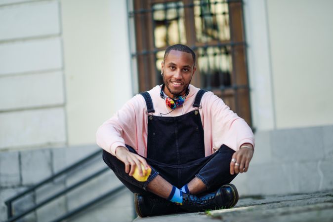 Calm male in overalls and boots sitting cross legged on outdoor steps with apple