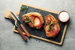 Board with steaks and cutlery on gray background. Grilled food
