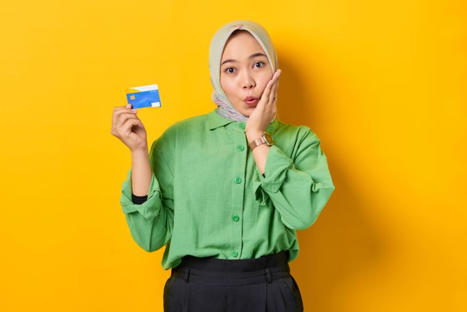 Muslim woman in headscarf and green blouse holding credit card and with hand to cheek in surprise