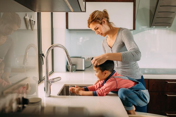 Small boy with mother washing hands under kitchen sink faucet