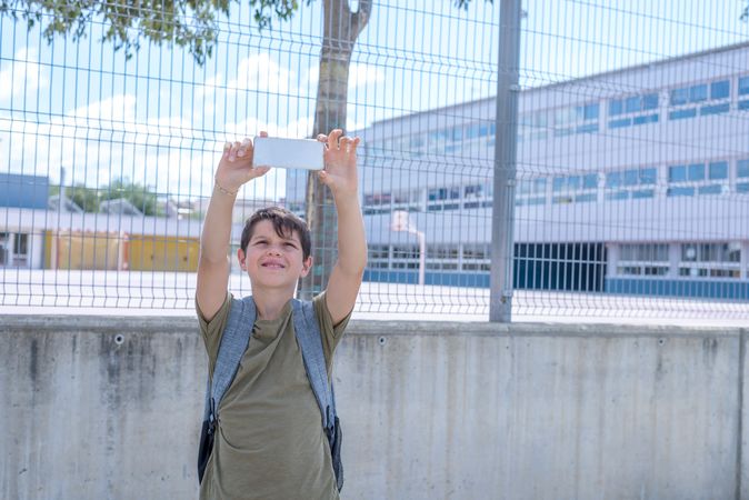Smiling teenage boy taking selfie in front of school fence