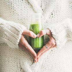 Woman’s hand making heart shape, holding green smoothie wearing sweater, square crop