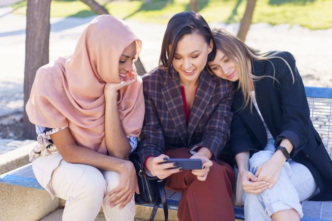 Smiling women sitting on outdoor park bench enjoying watching smartphone