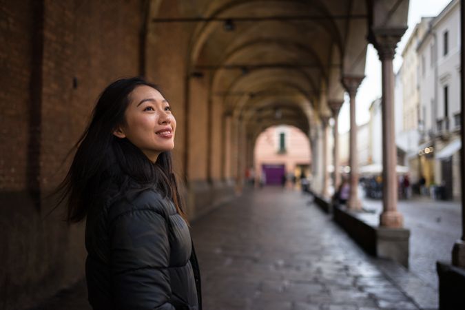 Joyful Asian female on vacation in warm clothing walking on old streets of city