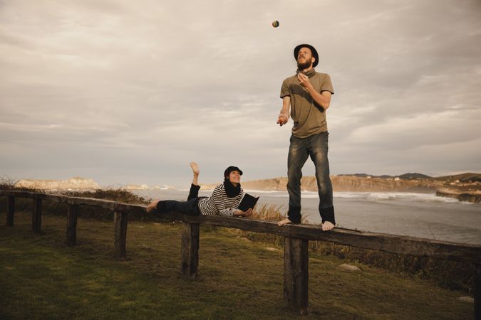 Man juggling balls on bench near woman with drum on shore