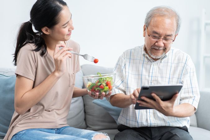 Woman feeding mature man salad at home while he uses tablet