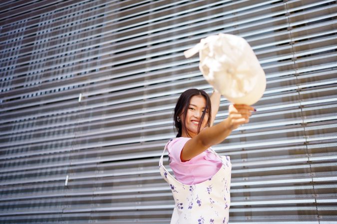 Smiling Chinese woman in floral coveralls throwing her handbag in front of metal wall