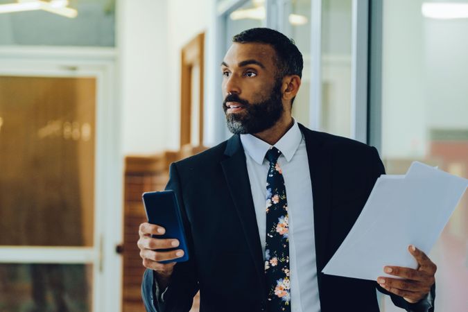 Serious businessman in suit and tie with papers standing in office while holding smart phone