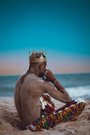 Older man wearing a crown and pants sitting on sand at the beach