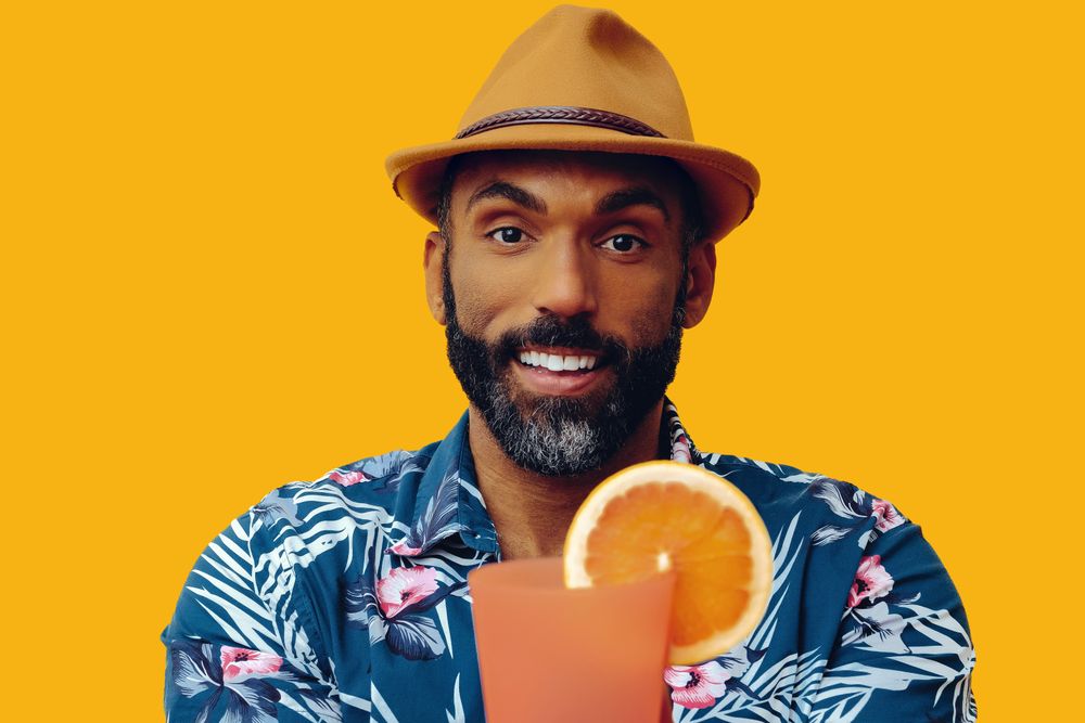Smiling male passing a cocktail to camera while wearing colorful shirt with yellow background