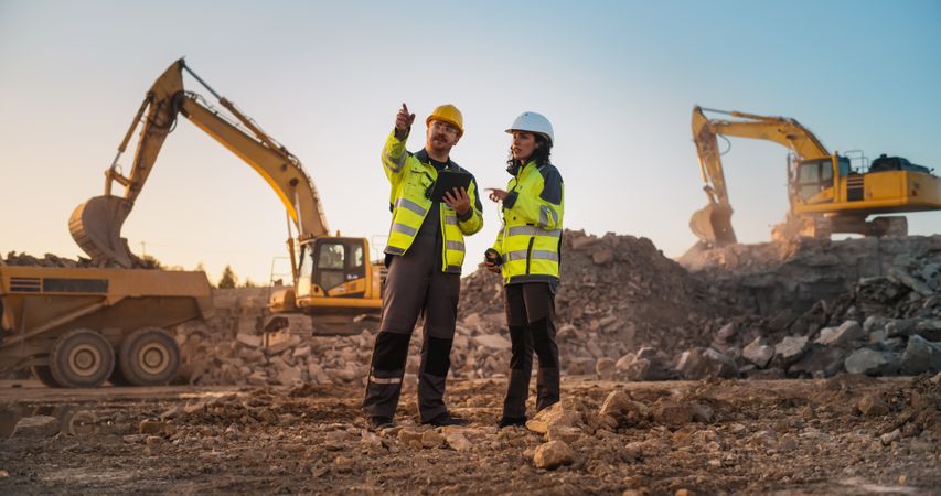 White Male Civil Engineer Talking To Hispanic Female Inspector And Using Tablet On Construction Site of Apartment Complex. Real Estate Developers Discussing Business, Excavators Working.