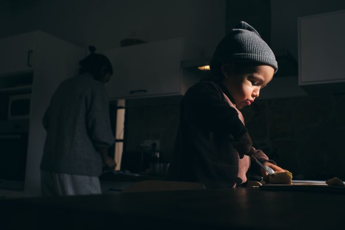 Focused little boy cutting banana on board near anonymous mother