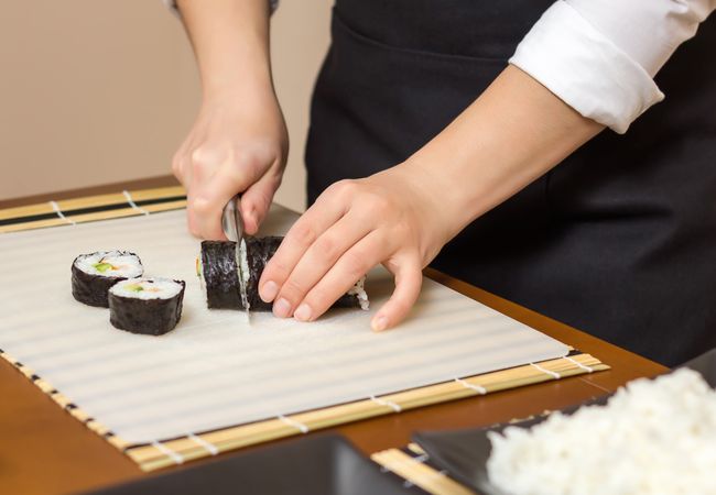 Female chef's hands cutting Japanese sushi rolls with rice, avocado and shrimps on nori seaweed sheet