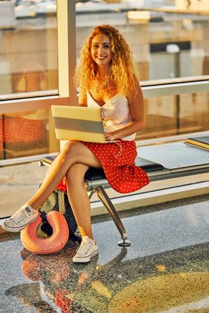Enthusiastic woman working on laptop sitting on bench in airport hall