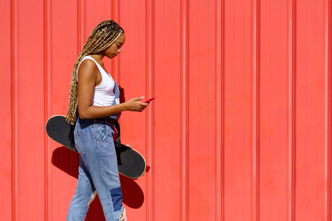 Woman walking in front of red wall with phone and skateboard