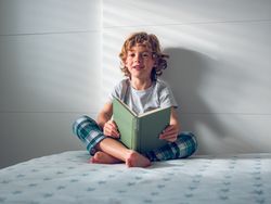 Cheerful curly blond boy reading book while sitting on bed