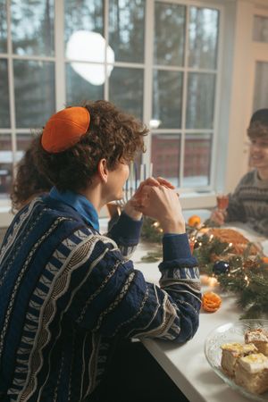 Back view of young man wearing kippah and sweater sitting at dinner table