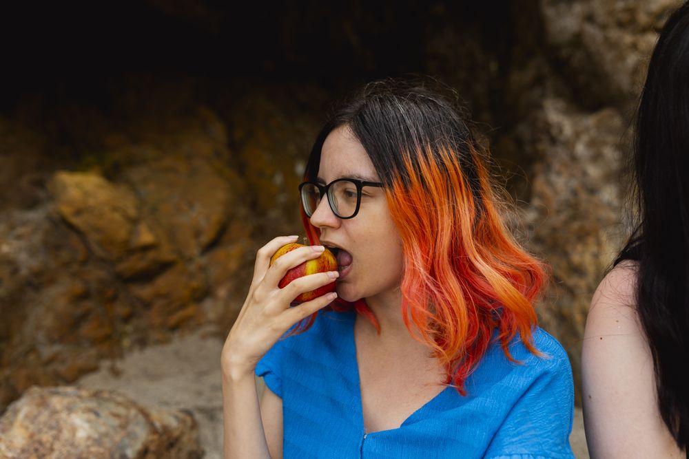 An orange-haired woman bites into an apple enjoying the outdoors
