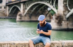 Bearded young man in casual wear using smartphone while walking on quay