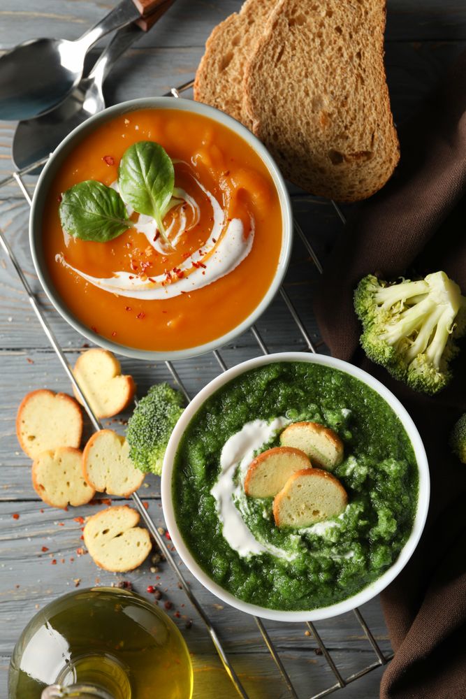 Two bowls of orange and green soup on wooden table with crackers and vegetables, vertical