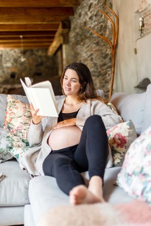Relaxed pregnant woman enjoying reading a book on her home sofa
