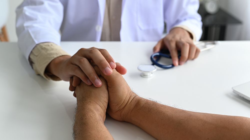 A medical professional in a lab gown with a stethoscope holds the patient hand to comfort