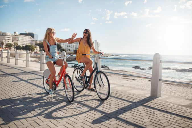 Two women friends riding bikes while smiling and giving high fives