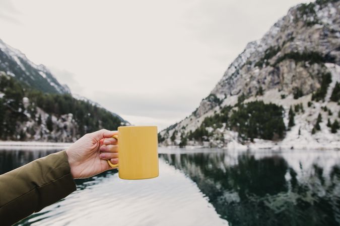 Person with cup near wonderful lake between hills in snow and cloudy sky