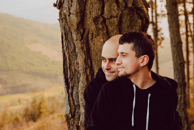 Smiling men hugging near tree in forest on hill