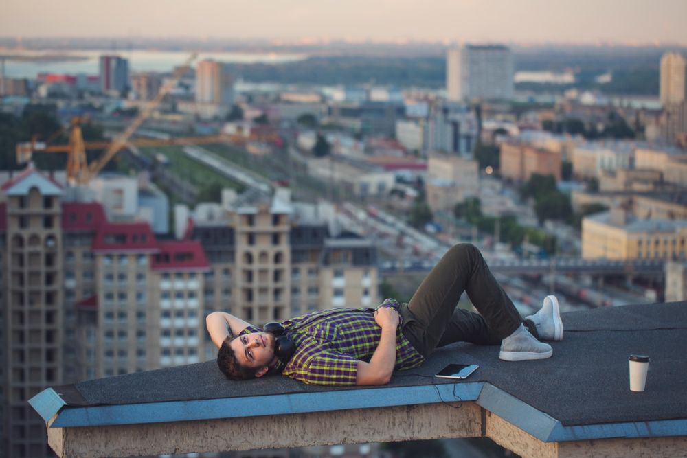 Male lying back looking at camera, with tablet, headphones and takeaway coffee cup on roof