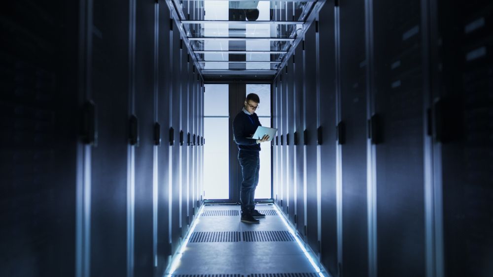 Male IT Engineer Works on a Laptop at the end of a Corridor in a Big Data Center. Rows of Rack Servers are Seen.