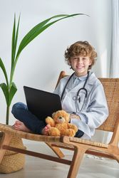 Cheerful boy doctor with laptop and soft bear on chair