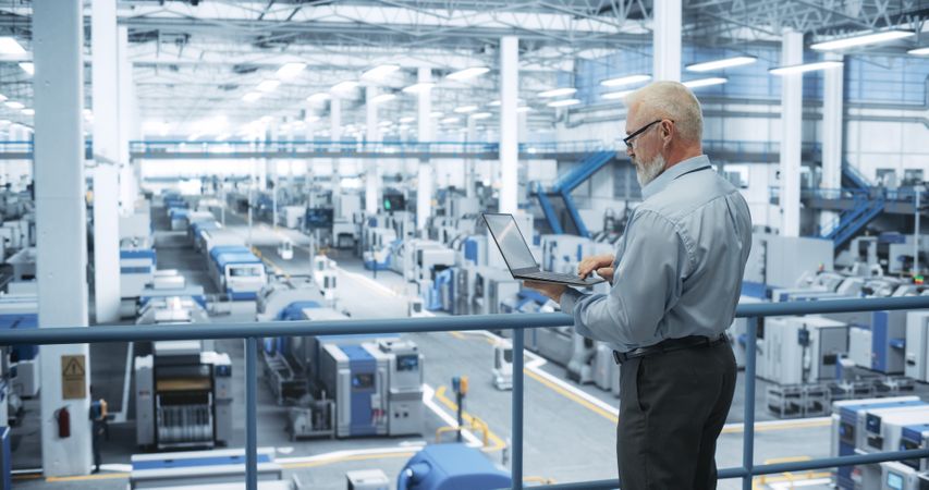Mature Factory Manager Working on Laptop at a Futuristic Industrial Manufacture with Autonomous AI Robots. Specialist Monitoring All the Data on a Computer, Checking Reports and Planning Workload
