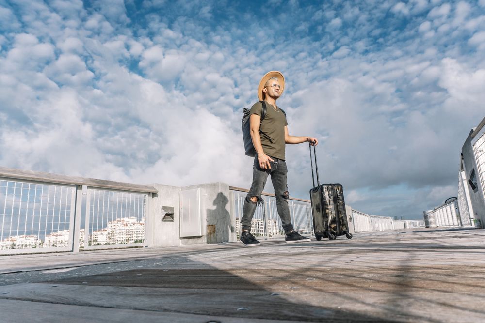 Full length side view serene tourist holding luggage while stands in a brown hat and ripped jeans