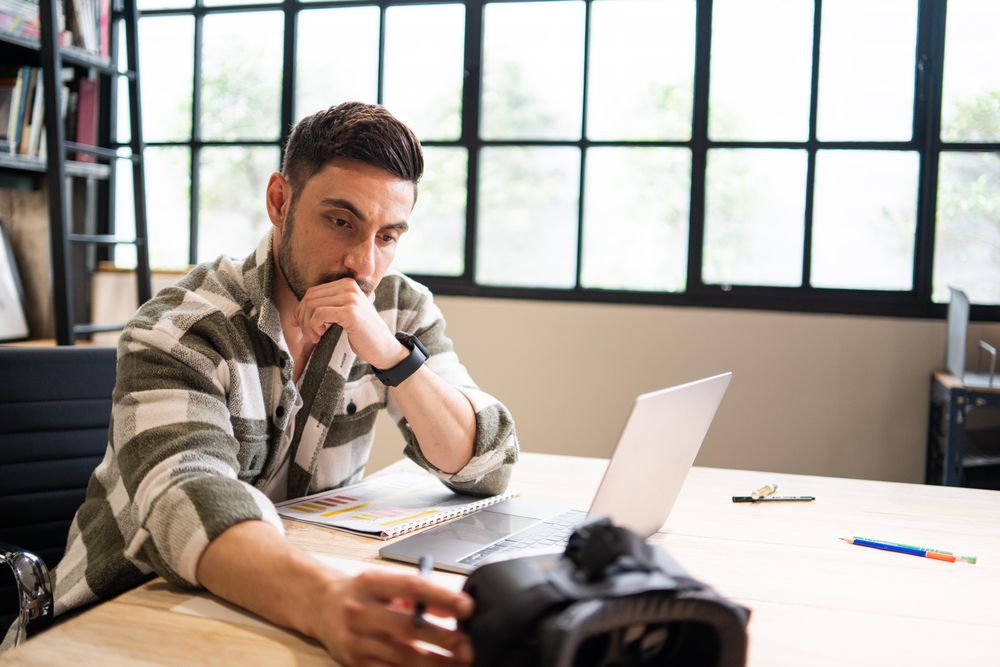 Serious young professional  looking at VR headset at work at a laptop in relaxed modern office
