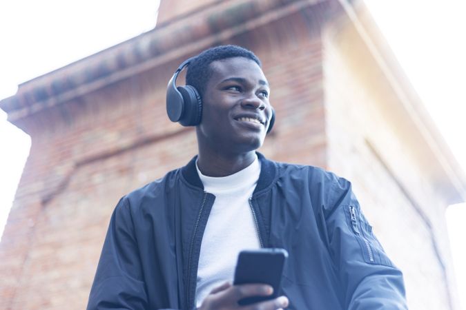 Smiling Black man sitting outside, wearing headphones and holding his phone