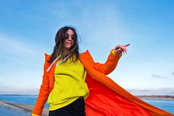 Trendy woman in sunglasses, orange coat, yellow hoodie and jeans in front of the coast
