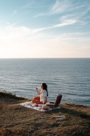 Woman with glass of drink on picnic mat looking at sea and mountains