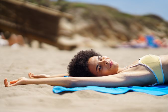 Female with curly hair in yellow bikini relaxing on blue towel on the sandy beach