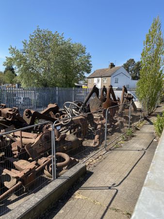 Cluster of rusted iron machine parts in a space surrounded by metal fence near house and trees