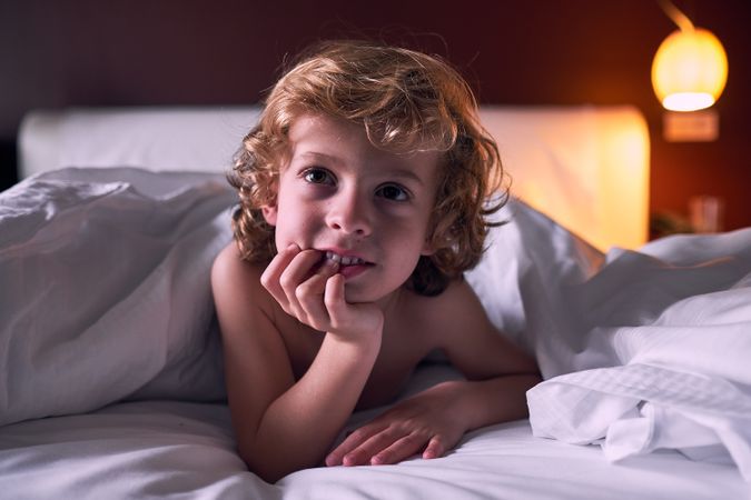 Curious little boy lying on bed under duvet