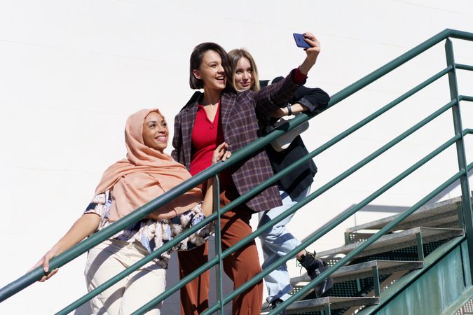 Multi-ethnic female friends, in stylish outfit and hijab taking selfie on outdoor staircase