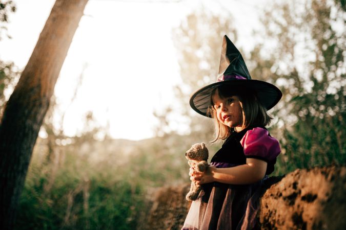 Girl in witch hat with teddy bear leaning on ridge in the forest