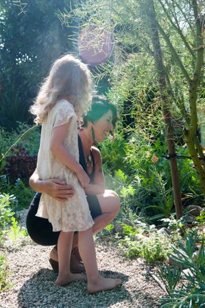 Smiling mother puts arm around  daughter on rock path exploring plants outside with lens flare