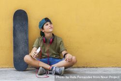 Front view of a teenage boy sitting on ground leaning on a yellow wall while holding a mobile phone bxAvMX